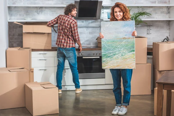 Pareja desembalaje cajas de cartón en el nuevo hogar, novia sosteniendo la pintura y mirando a la cámara - foto de stock