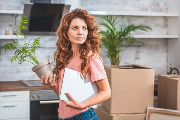 Beautiful woman with curly red hair holding potted plant and laptop at new home — Stock Photo