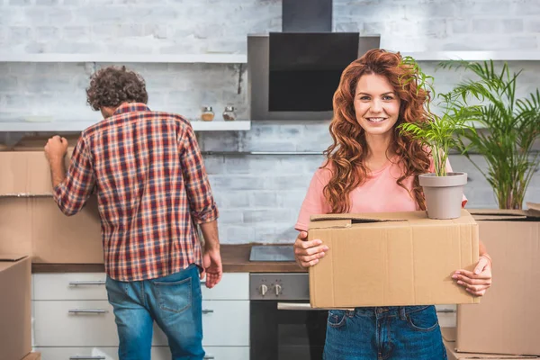 Paar packt Kartons im neuen Zuhause aus, Freundin hält Karton mit Pflanze in der Hand und blickt in die Kamera — Stockfoto