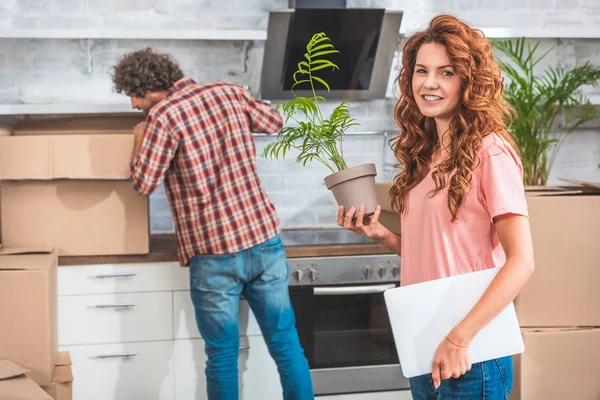 Pareja desembalaje cajas de cartón en el nuevo hogar, novia sosteniendo maceta planta y mirando a la cámara - foto de stock