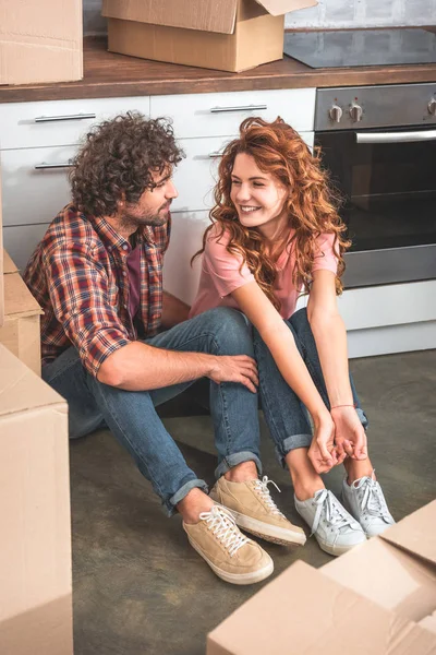 Vista de ángulo alto de alegre pareja sentada en el suelo cerca de cajas de cartón en la cocina nueva y mirándose entre sí - foto de stock