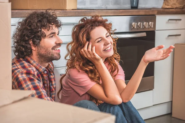 Sonriente pareja sentada en el suelo cerca de cajas de cartón en la cocina nueva y mirando hacia otro lado - foto de stock