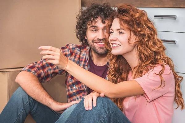 Cheerful couple sitting on floor near cardboard boxes in new kitchen, girlfriend gesturing and looking away — Stock Photo