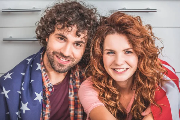 Portrait of smiling couple sitting wrapped in american flag and looking at camera — Stock Photo