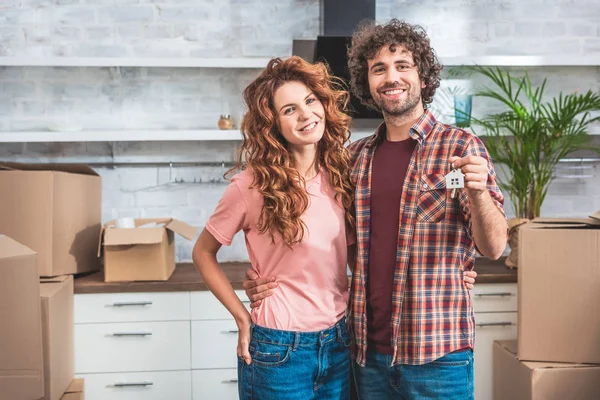 Coppia sorridente abbracciando e tenendo casa gingillo con le chiavi a casa nuova — Foto stock