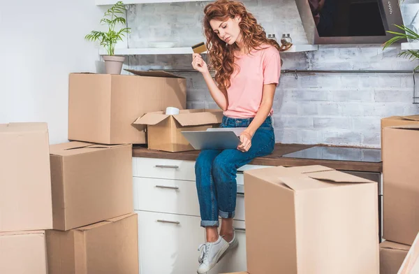 Attractive young woman holding credit card and using laptop while relocating in new apartment — Stock Photo