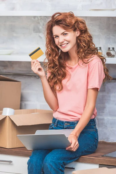 Atractiva mujer joven sosteniendo la tarjeta de crédito y sonriendo a la cámara mientras usa el ordenador portátil en el nuevo apartamento - foto de stock