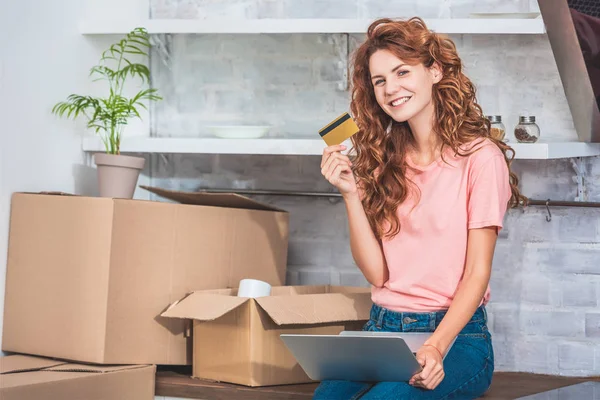 Hermosa mujer joven feliz sosteniendo la tarjeta de crédito y sonriendo a la cámara mientras se utiliza el ordenador portátil en el nuevo apartamento - foto de stock