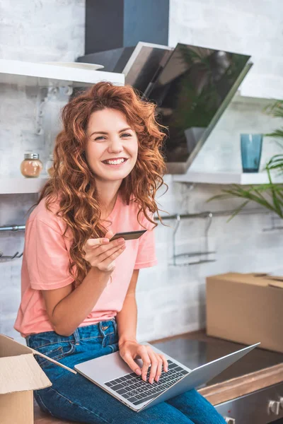 Jeune femme heureuse tenant la carte de crédit et utilisant un ordinateur portable dans un nouvel appartement — Photo de stock