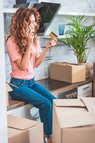 Happy young woman holding credit card and talking by smartphone while relocating in new apartment — Stock Photo