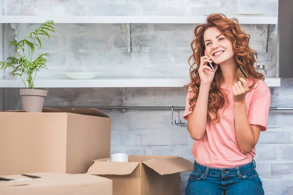 Mujer joven feliz sosteniendo tarjeta de crédito y hablando por teléfono inteligente durante la reubicación - foto de stock