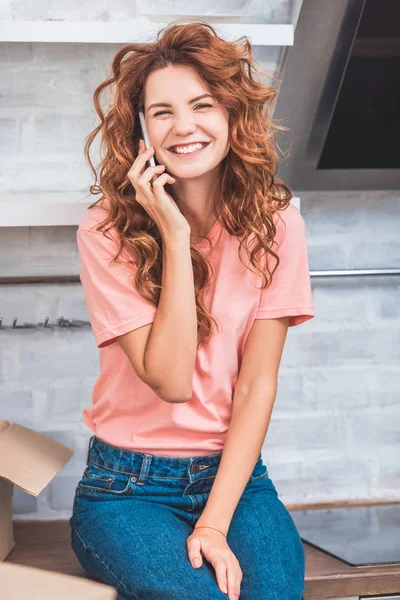 Feliz joven mujer hablando por teléfono inteligente y sonriendo a la cámara mientras se muda a casa - foto de stock