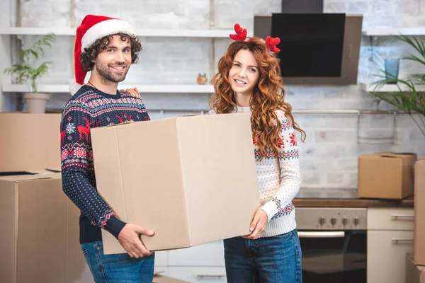 Feliz joven pareja sosteniendo caja de cartón y sonriendo a la cámara mientras se muda en un nuevo apartamento en Navidad - foto de stock