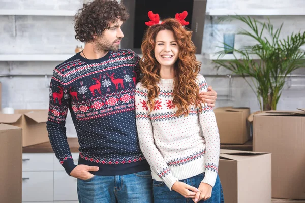 Young man looking at smiling girlfriend in antlers headband while standing near cardboard boxes in new apartment — Stock Photo