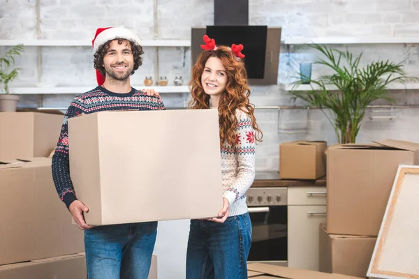 Happy young couple holding cardboard box and smiling at camera while relocating in new home at christmastime — Stock Photo
