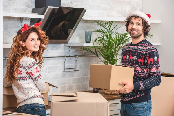 Glückliches junges Paar lächelt in die Kamera und packt zur Weihnachtszeit Pappkartons im neuen Zuhause aus — Stockfoto