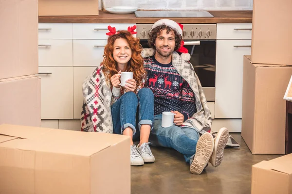 Felice giovane coppia che tiene le tazze e sorride alla macchina fotografica mentre si siede tra scatole di cartone a Natale — Foto stock