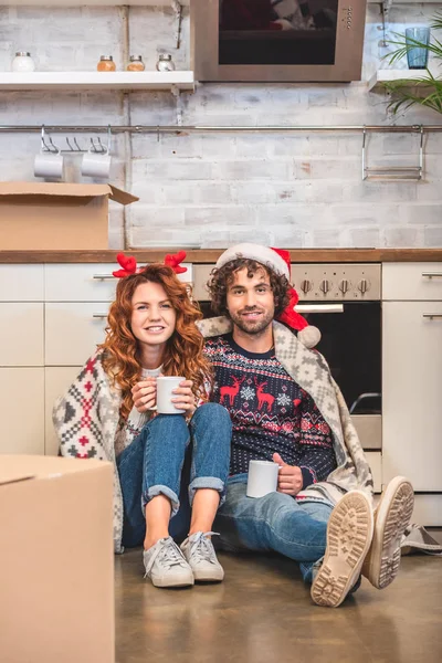 Feliz joven pareja sosteniendo tazas y sonriendo a la cámara mientras está sentado en un nuevo apartamento en Navidad - foto de stock