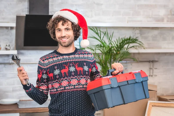 Felice giovane uomo in cappello di Babbo Natale in possesso di cassetta degli attrezzi e sorridente alla fotocamera nel nuovo appartamento — Foto stock