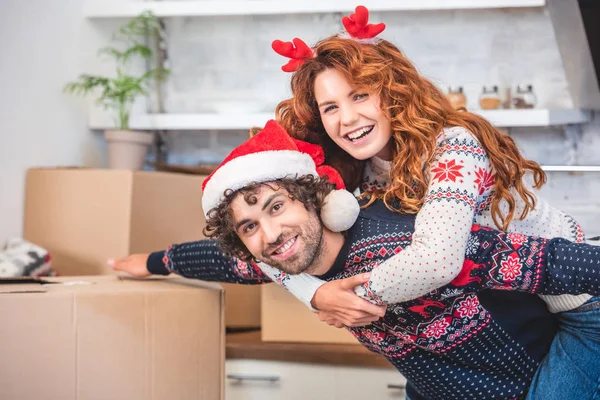 Happy young couple piggybacking and smiling at camera while relocating at christmastime — Stock Photo