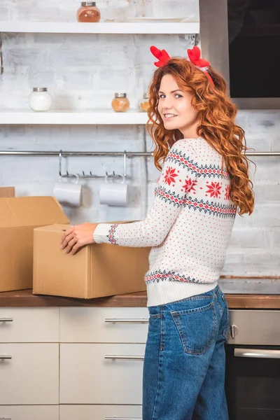 Beautiful young woman in antlers headband holding cardboard box and smiling at camera while relocating in new apartment — Stock Photo