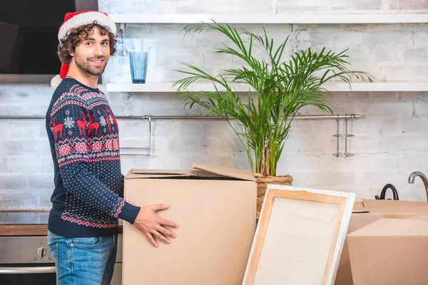 Beau jeune homme heureux dans santa chapeau tenant boîte en carton et souriant à la caméra lors de la relocalisation — Photo de stock