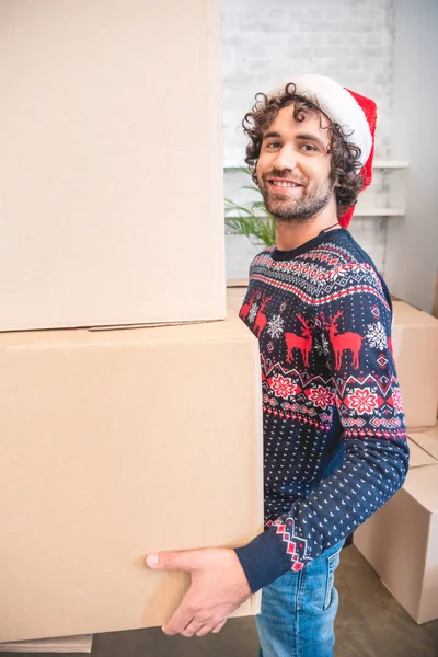 Счастливый молодой человек в шляпе санта, держащий картонные коробки и улыбающийся на камеру — стоковое фото