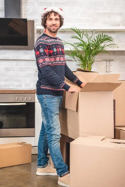 Handsome young man in santa hat unpacking cardboard boxes and smiling at camera — Stock Photo