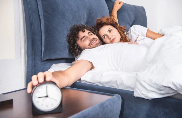 Happy young couple waking up together, man reaching to alarm clock — Stock Photo