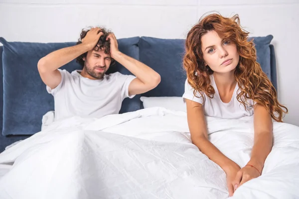 Jovem mulher sentada na cama e olhando para a câmera, enquanto o homem que sofre de dor de cabeça atrás — Fotografia de Stock