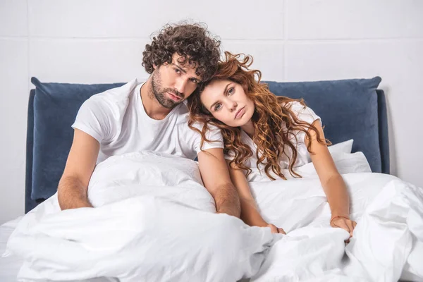 Junges Paar leidet unter Kopfschmerzen, sitzt auf dem Bett und blickt in die Kamera — Stockfoto