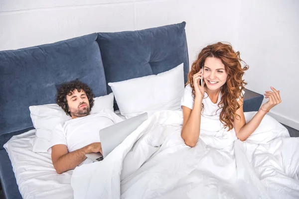 Vue grand angle de femme souriante parlant par smartphone et l'homme en utilisant un ordinateur portable sur le lit — Stock Photo