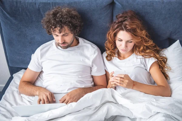 Vista ad alto angolo di giovane coppia utilizzando laptop e smartphone a letto — Foto stock