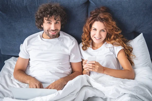 Vista de ángulo alto de feliz pareja joven utilizando dispositivos digitales y sonriendo a la cámara mientras está acostado en la cama - foto de stock