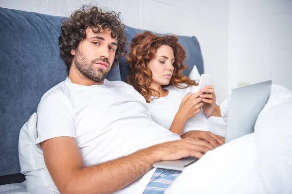 Young couple using laptop and smartphone in bed — Stock Photo