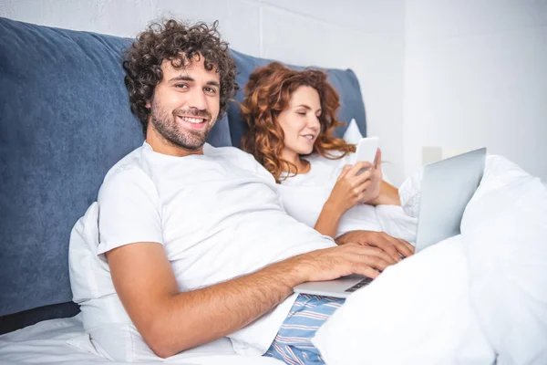 Feliz pareja joven utilizando dispositivos digitales en la cama - foto de stock