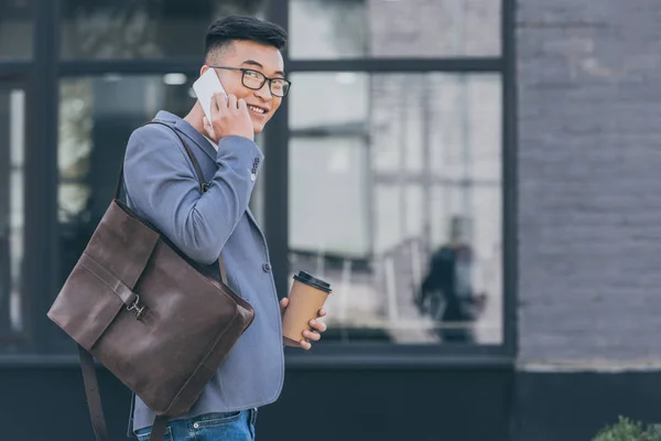 Guapo asiático hombre con cuero mochila y café para ir a hablar en smartphone - foto de stock