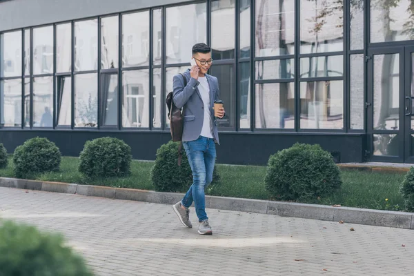 Heureux asiatique homme avec café pour aller parler sur smartphone tout en marchant sur la rue — Photo de stock