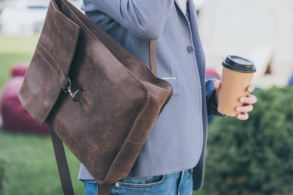 Vista recortada del hombre con mochila de cuero que sostiene la taza de café desechable - foto de stock