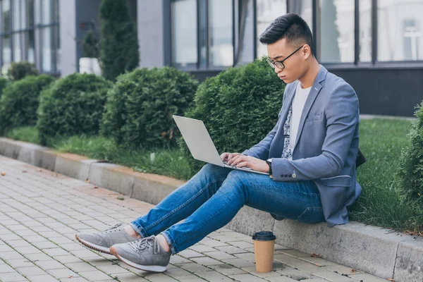 Seguro asiático freelancer trabajando en portátil mientras está sentado en la acera con taza de café desechable - foto de stock