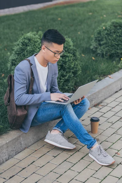 Красивый азиатский мужчина телеработает с ноутбуком, сидя на тротуаре — стоковое фото