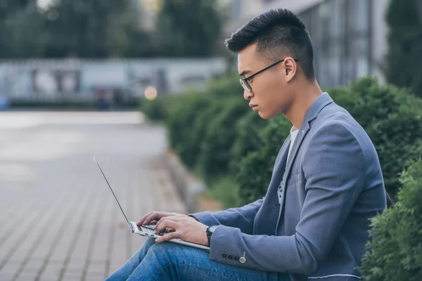 Confident asian freelancer in eyeglasses working on laptop while sitting on sidewalk — Stock Photo