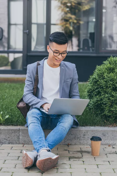 Усміхнений азіатський телефонний працівник використовує ноутбук, сидячи на тротуарі з кавою, щоб піти — стокове фото