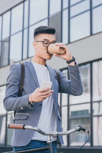 Asian man drinking coffee to go and looking at smartphone near bicycle in city — Stock Photo