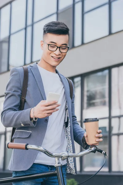 Happy asian man with coffee to go looking at smartphone and standing near bike — Stock Photo