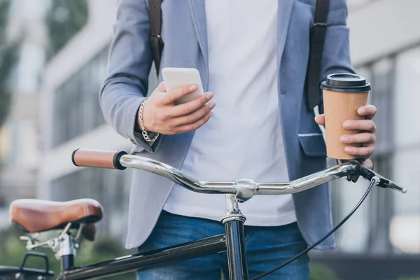 Vista parziale dell'uomo in possesso di tazza monouso di caffè e utilizzando smartphone vicino alla bicicletta — Stock Photo