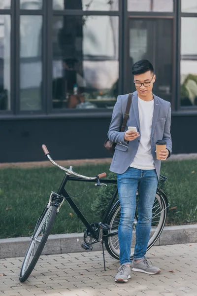 Стильний азіатський чоловік з одноразовою чашкою за допомогою смартфона і стоячи біля велосипеда — стокове фото