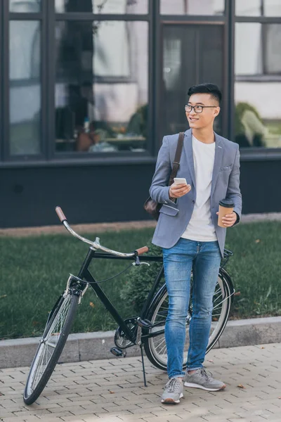 Sorrindo asiático homem com café para ir usando smartphone e de pé perto de bicicleta — Fotografia de Stock