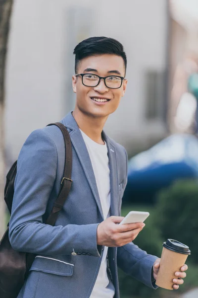 Усміхнений азіатський чоловік тримає одноразову чашку кави і використовує смартфон — стокове фото