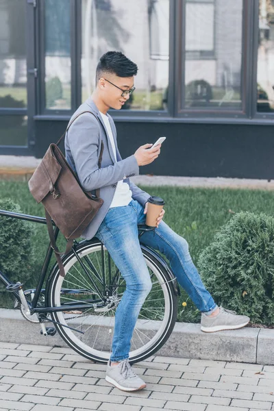 Handsome asian man with leather backpack and coffee to go using smartphone and sitting on bike — Stock Photo